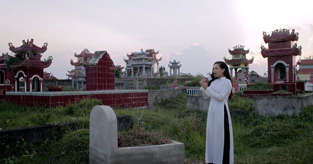 Still from the film "Memoryland" by Kim Quy Bui. We see a woman dressed in white in a graveyard, singing in front of a grave with a karaoke microphone. 