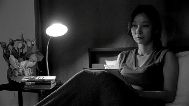 Black and white image of a woman sitting on the back of a bed. She has a notebook on her lap. To her left is a bedside table with a lamp, flowers and books. 