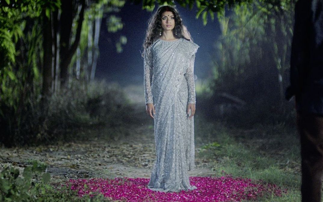 Filmstill aus "In the Belly of a Tiger" von Siddartha Jatla. Zu sehen ist eine Frau in einem glitzernden silbernen Kleid auf dem Lande. Sie steht auf Blumen.