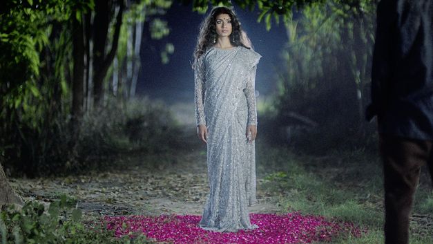 Film still from "In the Belly of a Tiger" by Siddartha Jatla. It shows a woman in a silver sparkly dress in the countryside. She stands on flowers.