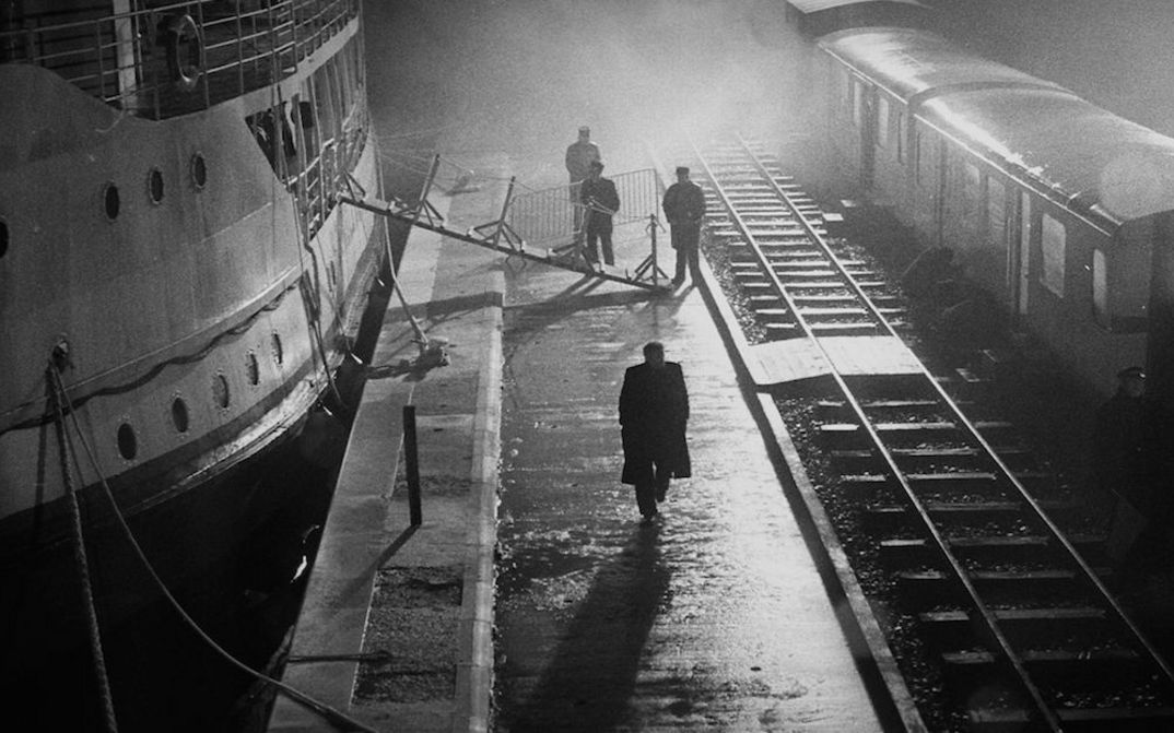 Filmstill aus MAN FROM LONDON: Nächtliche Aufnahme mit starken Lichtkontrasten. Zwischen einer Schiffsanlegestelle und Bahngleisen sind vereinzelte Menschen zu sehen.