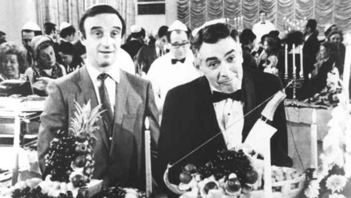 Film still from THE PLOT AGAINST HARRY: Two men at a party are standing in front of a richly laid table. 