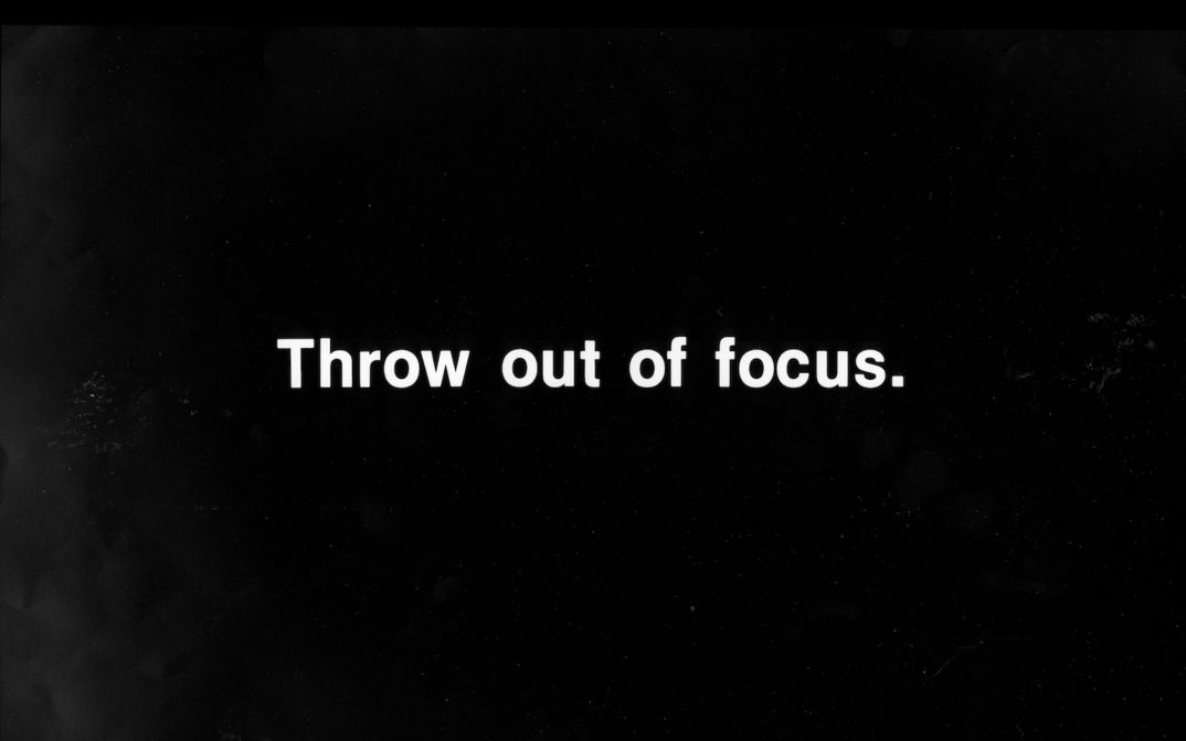 Film still from PROJECTION INSTRUCIONS: In white lettering on a black background it says: Throw out of focus.