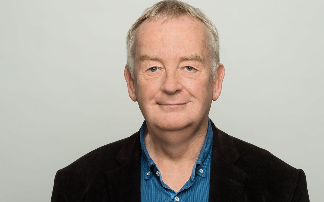 Porträtbild von Ralf Schenk. Er trägt ein Jacket und blaues Hemd und lächelt in die Kamera.