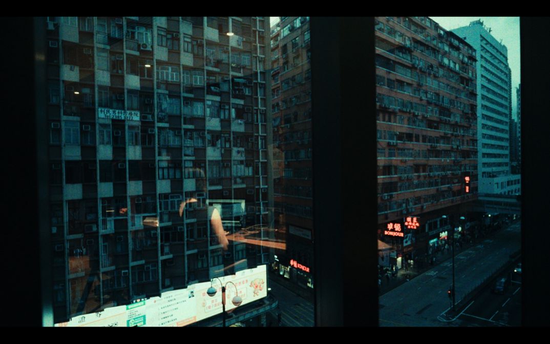 Film still from Elysa Wendi and Wai Shing Lee’s film “Room 404”. The view from a window on blocks and blocks of apartments, and in the reflection of the window, a person sits in front of what seems like a computer screen.