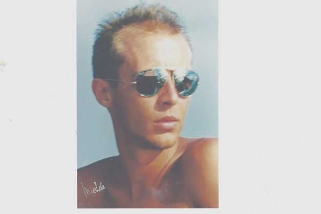 Porträt eines jungen Mannes mit Sonnenbrille, nackter Oberkörper, neutraler Hintergrund.