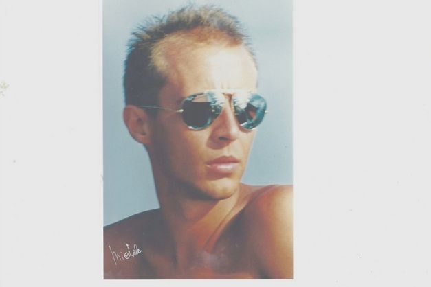 Porträt eines jungen Mannes mit Sonnenbrille, nackter Oberkörper, neutraler Hintergrund.