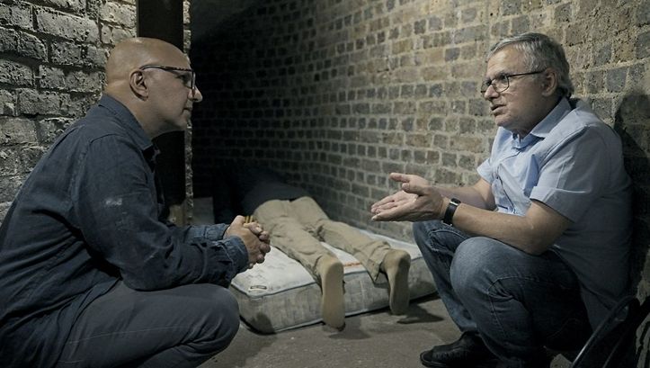 Still from the film "Jaii keh khoda nist" by Tamadon Mehran. Two men are crouching between two walls, in conversation. A lifeless body is lying on a mattress in the background.