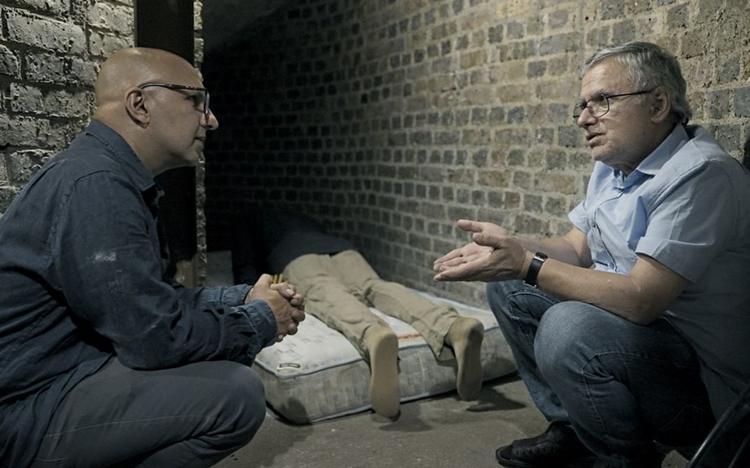 Filmstill aus dem Film "Jaii keh khoda nist" von Tamadon Mehran. Zwei Männer hocken zwischen zwei Wänden und unterhalten sich. Im Hintergrund liegt ein lebloser Körper auf einer Matratze.