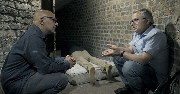 Still from the film "Jaii keh khoda nist" by Tamadon Mehran. Two men are crouching between two walls, in conversation. A lifeless body is lying on a mattress in the background.