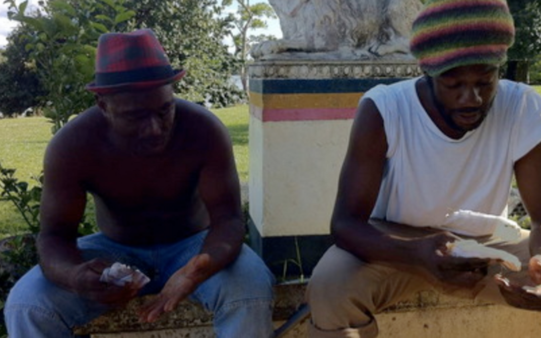 Film still from ALLER AU DIABLE: Two men are sitting next to each other on a small wall outside.