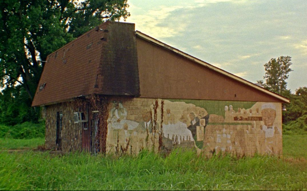Film still from Theresa Delsoin, Lisa Malloy, JP Sniadecki, Ray Whitaker’s film “On the Battlefield”. A small house in the middle of an open field with a mural on one side. 
