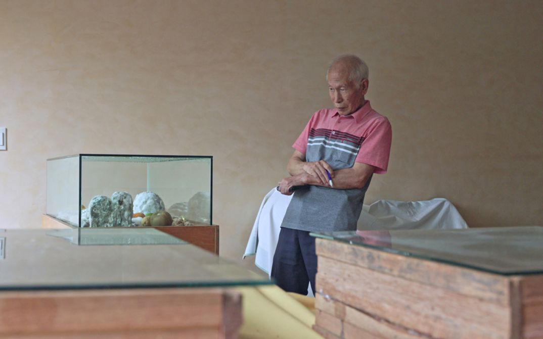 Film still from Jeamin Cha’s film “Photosynthesizing Dead in Warehouse”. A middle-aged man observes the decomposition of organic elements placed under a glass display case.