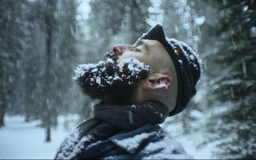 Film still from THE DAMNED: A man stands in a snow-covered forest. He is stretching his face towards the sky, his beard covered in snowflakes.