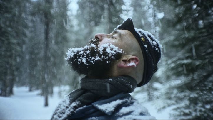 Film still from THE DAMNED: A man stands in a snow-covered forest. He is stretching his face towards the sky, his beard covered in snowflakes.