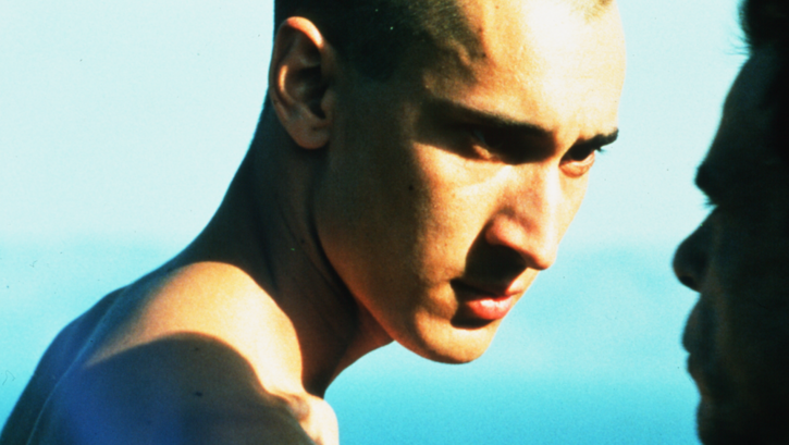 Filmstill aus BEAU TRAVAIL: Zwei Männer mit nacktem Oberkörper schauen sich intensiv in die Augen. Im Hintergrund ist das blaue Meer zu sehen.