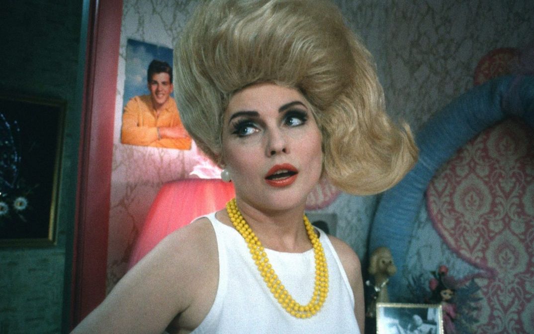 Filmstill aus HAIRSPRAY: Eine Frau mit blonden auftoupierten Haaren und gelber Perlenkette schaut nach links und steht in einem Zimmer mit auffälliger Wandtapete und Postern im Hintergrund.