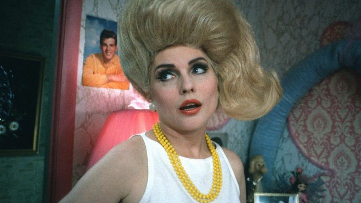 Filmstill aus HAIRSPRAY: Eine Frau mit blonden auftoupierten Haaren und gelber Perlenkette schaut nach links und steht in einem Zimmer mit auffälliger Wandtapete und Postern im Hintergrund.