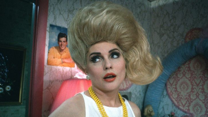 Filmstill from HAIRSPRAY: A woman with tousled blonde hair and a yellow pearl necklace looks to the left and stands in a room with eye-catching wallpaper and posters in the background.