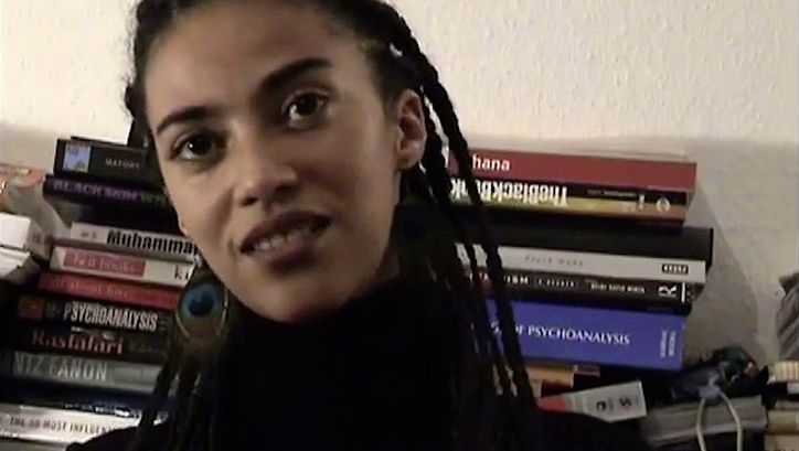 Still from the film "The Maji-Maji Readings" by Ricardo Barcallo. Close up of a women sitting in front of books