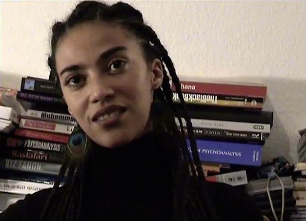 Still from the film "The Maji-Maji Readings" by Ricardo Barcallo. Close up of a women sitting in front of books
