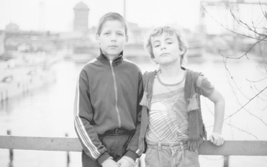Filmstill aus DDR – Ohne Titel: Zwei Jungen stehen auf einer Brücke und blicken in die Kamera. Im Hintergrund ist verschwommen eine Stadt zu sehen.