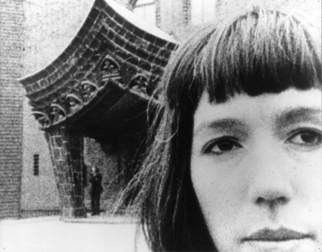 Film still from JOURNEYS FROM BERLIN/1971: We see the large face of a woman, in the background is the entrance to a building.