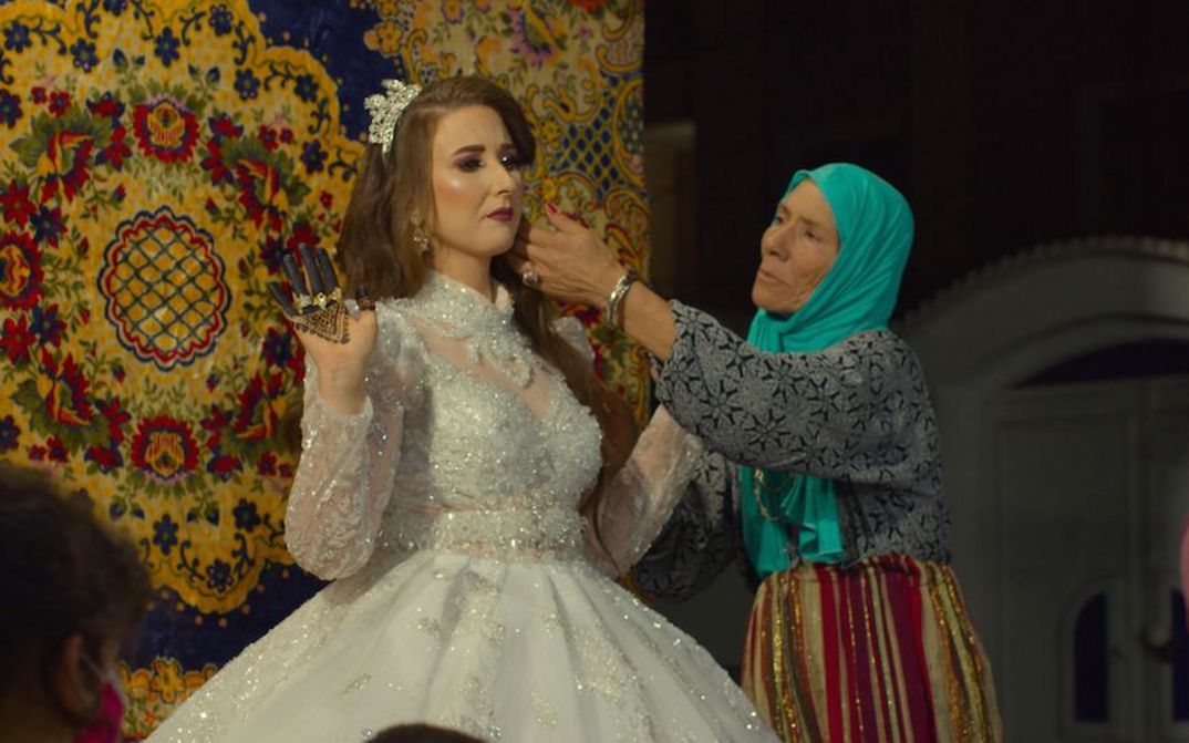 Filmstill aus MACHTAT: Eine ältere Frau mit türkisem Kopftuch richtet die Haare einer Braut.