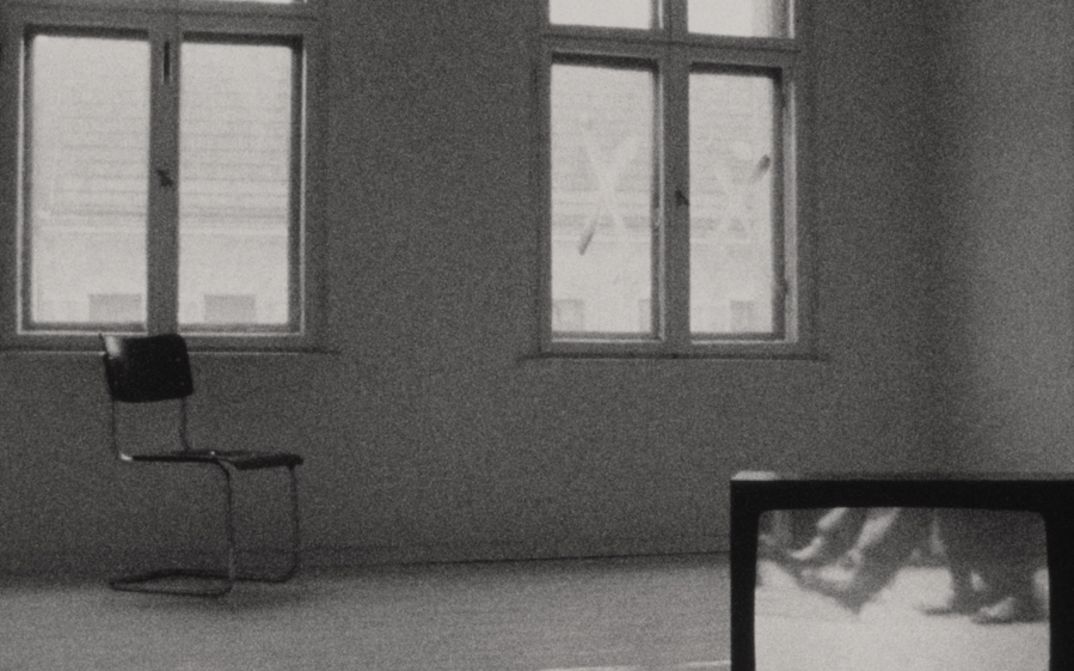 Film still from PERSONA NON GRATA: A view of a room that is empty except for a chair standing under one of the two windows and a television sitting on the floor in the foreground.
