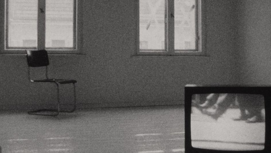 Filmstill aus PERSONA NON GRATA: Ansicht eines Raumes, der leer ist bis auf einen Stuhel, der unter einem der beiden Fenster steht, und einem Fernseher, der im Vordergrund auf dem Boden steht.