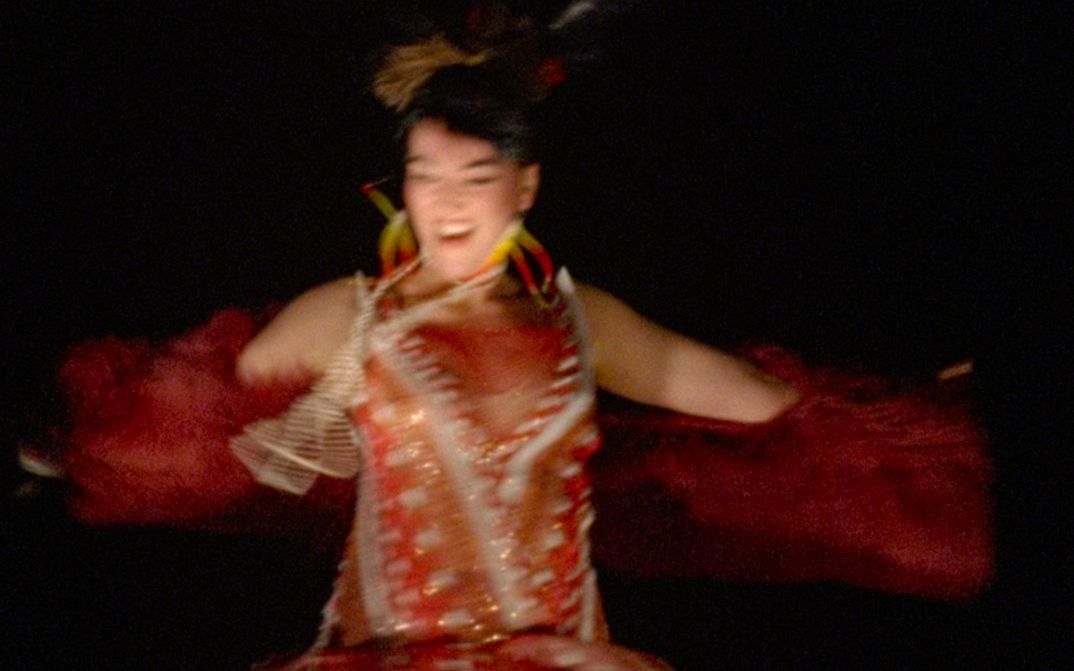 Film still from Caroline Monnet’s film “Pidikwe”. A person appears to be dancing or spinning, with arms spread wide and a radiant expression. The blurred image captures the rapid movement of the moment. They are wearing a contemporary creation that dialogues with Native American clothing heritage, blending red and golden tones, with geometric patterns and fringes that wave with the movement.