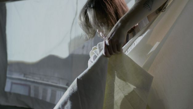 Filmstill from the film "The Song of the Shirt" by Kerstin Schroedinger. You see a person stretching out a white fabric.