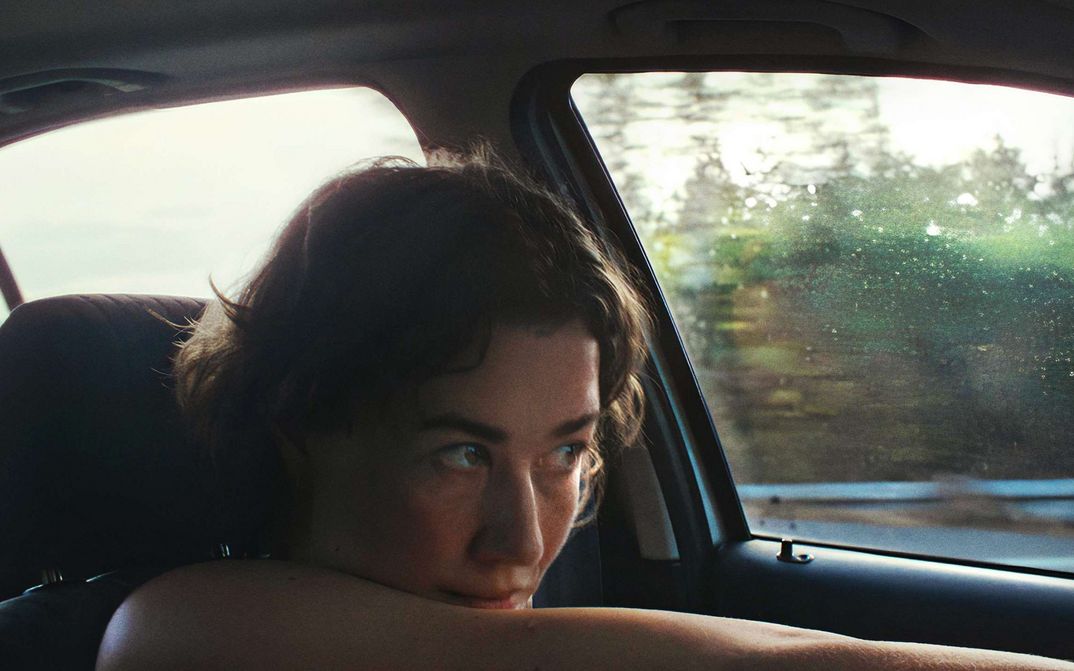 Filmstill aus IVO: Eine Frau sitzt im Auto, durch das Fenster sieht man die Landschaft vorbeiziehen.