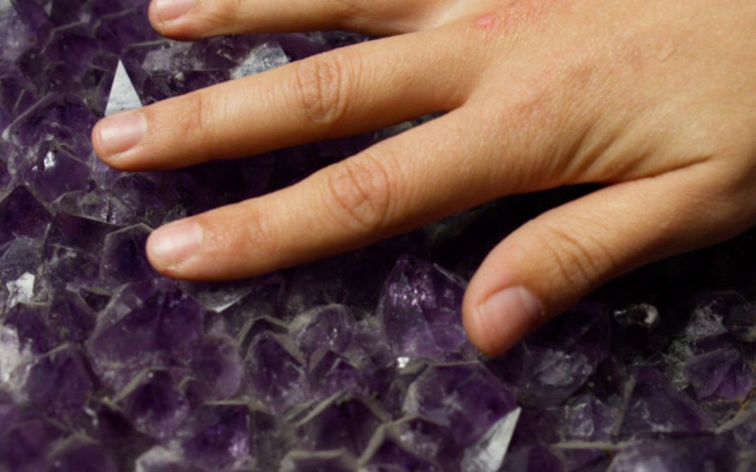 Hand berührt violette Kristalle mit scharfkantiger, unregelmäßiger Oberfläche.