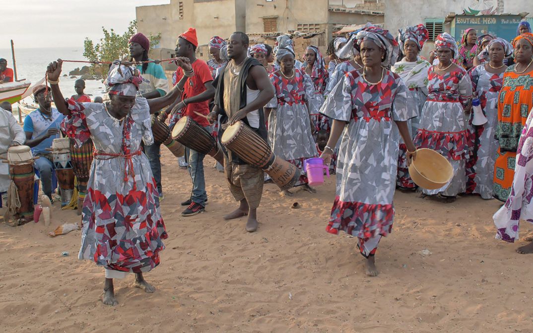 Still from the film "AI: African Intelligence" by Manthia Diawara. Drummers and a group of colourfully dressed dancers in a village square.