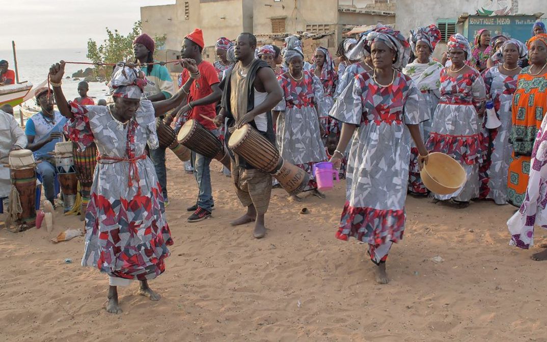 Filmstill aus dem Film „AI: African Intelligence“ von Manthia Diawara. Eine Gruppe von Menschen mit Trommeln und Menschen in bunten Gewändern auf einem Platz zwischen Häusern mit dem Meer im Hintergrund.