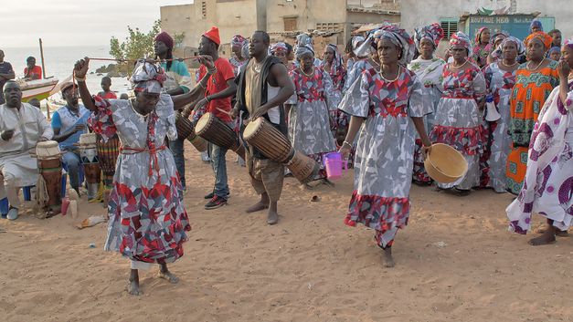 Still from the film "AI: African Intelligence" by Manthia Diawara. Drummers and a group of colourfully dressed dancers in a village square.