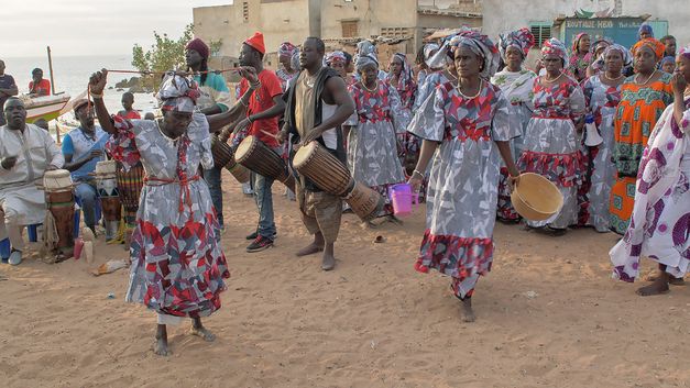 Filmstill aus dem Film „AI: African Intelligence“ von Manthia Diawara. Eine Gruppe von Menschen mit Trommeln und Menschen in bunten Gewändern auf einem Platz zwischen Häusern mit dem Meer im Hintergrund.