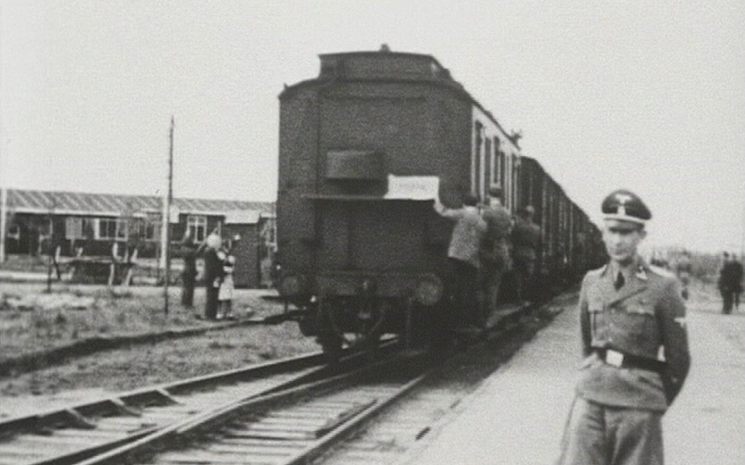 Film still from AUFSCHUB: You can see a historical photograph. An SS man stands in front of a departing train, other people can be seen in the background.