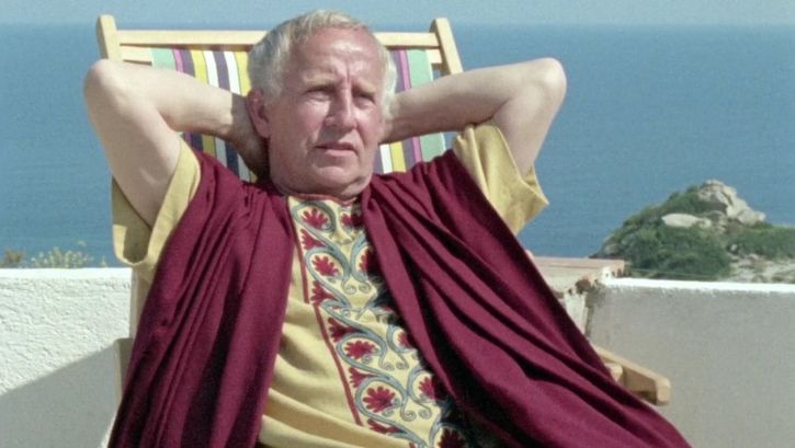 Filmstill aus GESCHICHTSUNTERRICHT: Ein Mann sitzt in einem römischen Kostüm in einem Liegestuhl vor dem Meer.