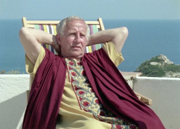 Film still from GESCHICHTSUNTERRICHT: A man in a Roman costume sits in a deck chair in front of the sea.