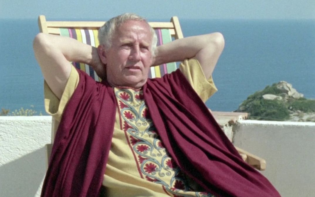 Filmstill aus GESCHICHTSUNTERRICHT: Ein Mann sitzt in einem römischen Kostüm in einem Liegestuhl vor dem Meer.
