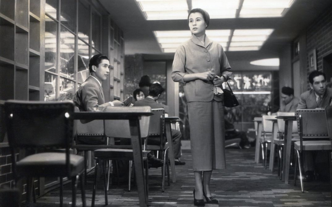 Filmstill aus KOIBUMI: Eine Frau steht in einem Lokal und blickt suchend um sich.