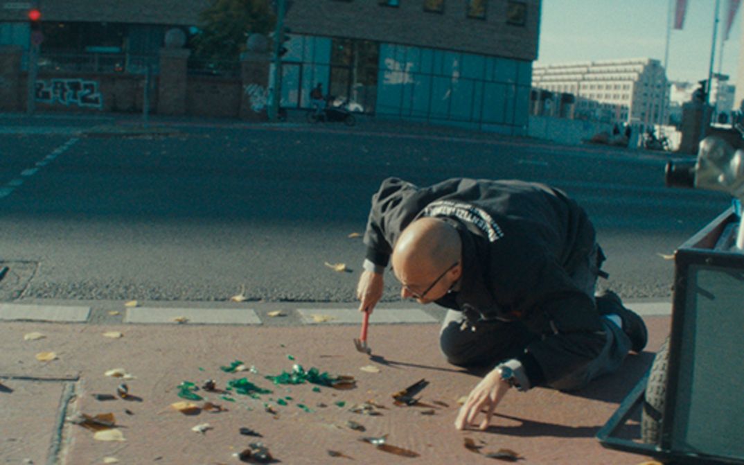 Film still from THE ART OF AUTHENTICITY: A man kneels on a sidewalk and examines the shards of a bottle. He holds a hammer in his hand.