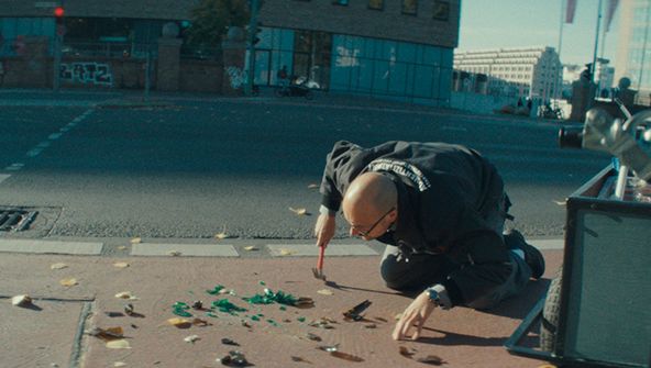 Filmstill aus THE ART OF AUTHENTICITY: Ein Mann kniet auf einem Gehweg und begutachtet die Scherben einer Flasche. In der Hand hält er einen Hammer.