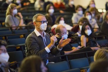 The cinema audience is wearing masks. A man is asking a question into a microphone.