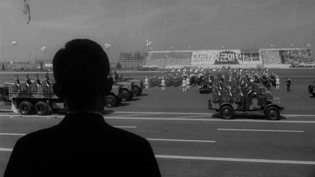 Film still from THE SENSE OF VIOLENCE by Mooyung Kim. The black and white image shows a military parade. In the front is the silhouette of a person.