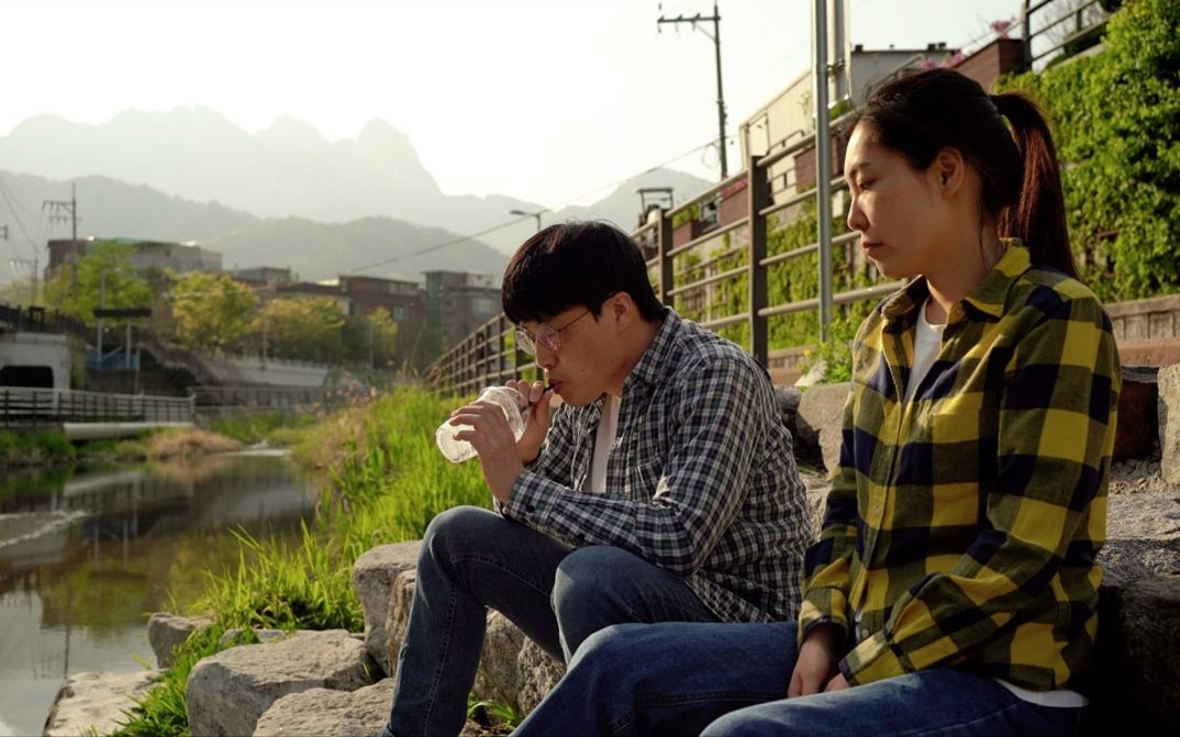 Filmstill aus „Hot in Day, Cold at Night“ von Park Song-yeol. An einem sonnigen Tag sitzen ein Mann und eine Frau an einem Bach. Der Mann trinkt etwas mit einem Strohhalm aus einem Plastikgefäß.