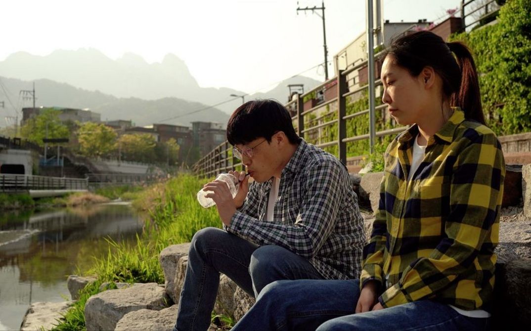 Filmstill aus „Hot in Day, Cold at Night“ von Park Song-yeol. An einem sonnigen Tag sitzen ein Mann und eine Frau an einem Bach. Der Mann trinkt etwas mit einem Strohhalm aus einem Plastikgefäß.