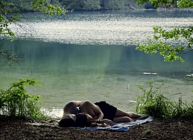 Filmstill aus „Afterwater“ von Dane Komljen. Ein Paar liegt aneinander geschmiegt am Seeufer. 